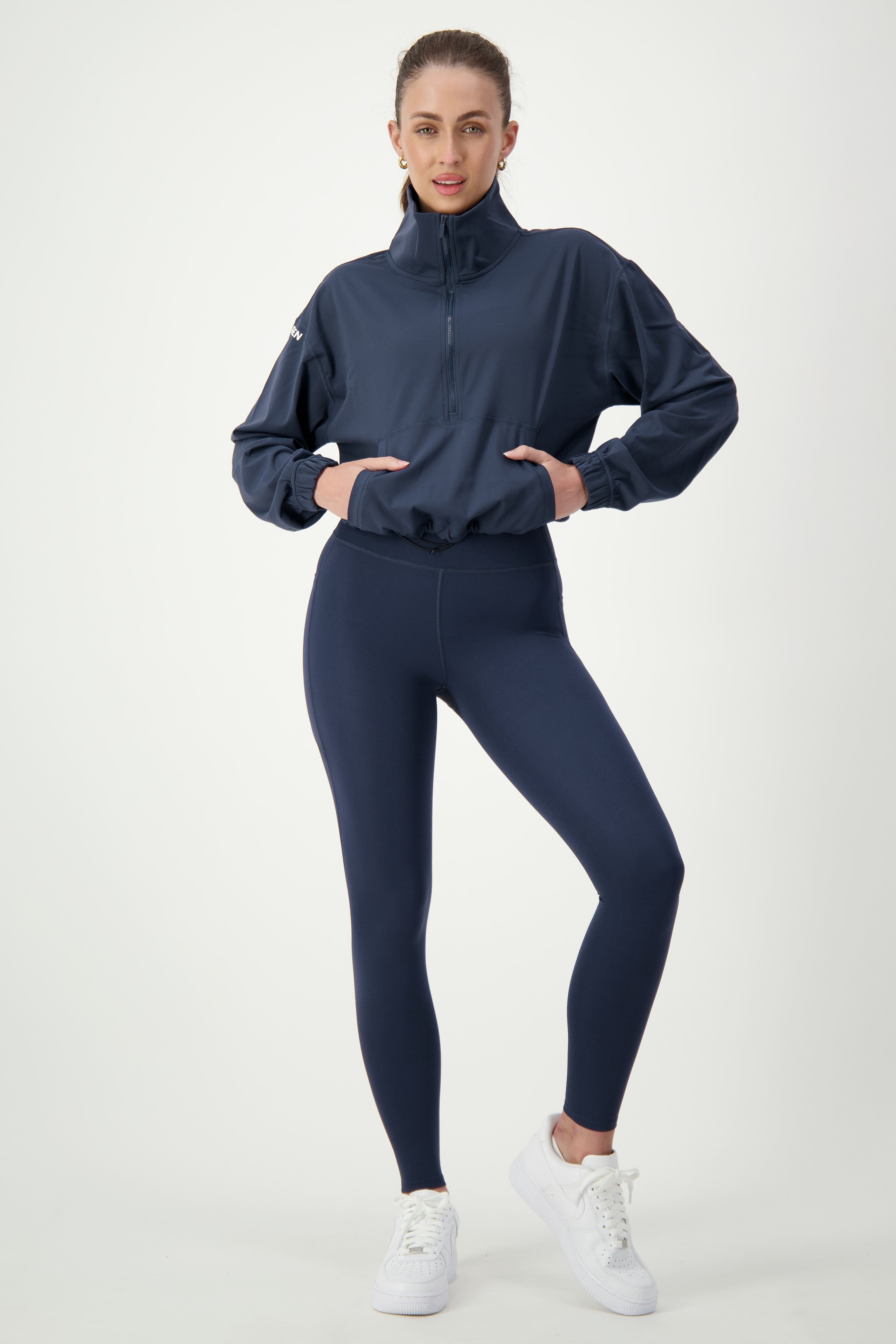 A person stands against a plain white background, wearing a dark blue Lara Half Zip Jacket (navy) by DRIVEN Gym Apparel and matching leggings that epitomize athleisure. They are also sporting white sneakers. The individual has one hand on their hip and a neutral expression.