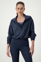 A woman with long brown hair tied back in a ponytail is wearing navy gym wear, with a Lara Half Zip Jacket (navy) by DRIVEN Gym Apparel partially open to reveal a top that reads "DRIVEN." She is also sporting matching navy leggings. The background is a plain, light gray color.