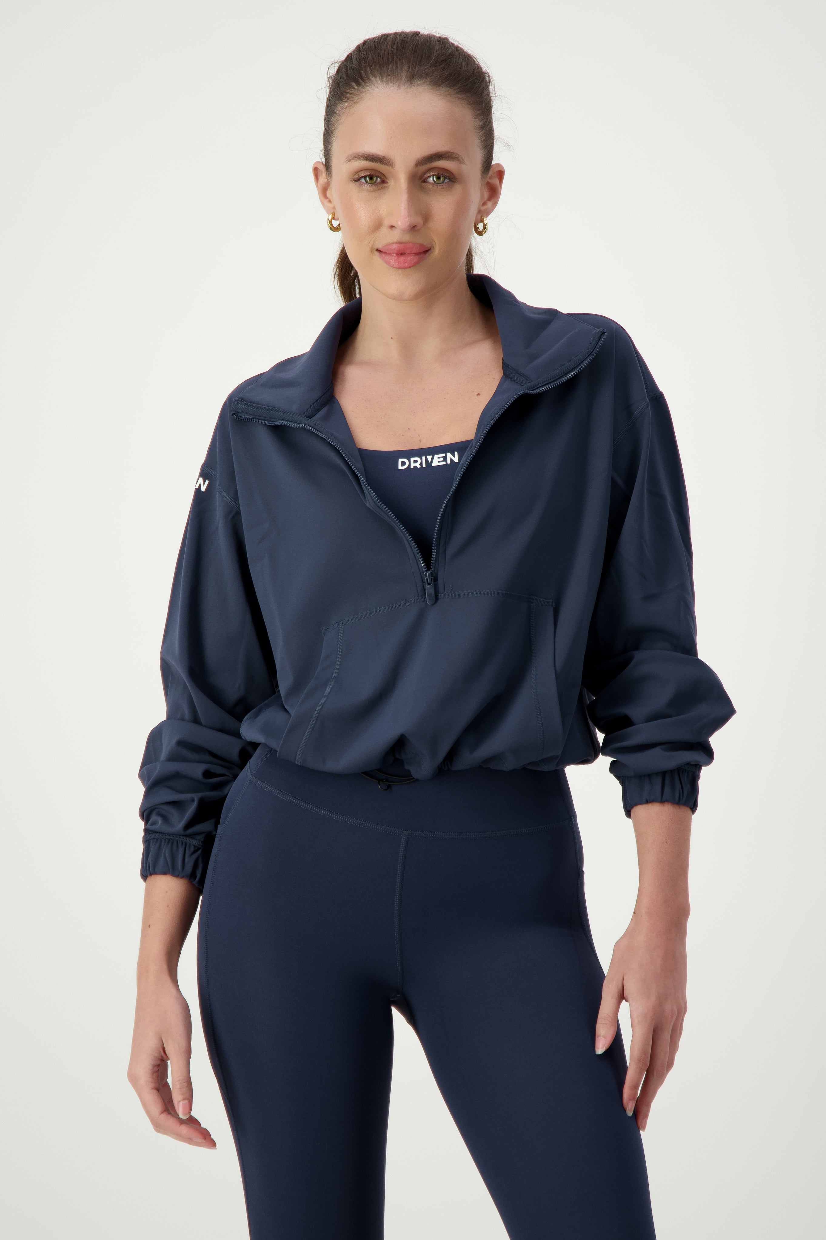 A woman with long brown hair tied back in a ponytail is wearing navy gym wear, with a Lara Half Zip Jacket (navy) by DRIVEN Gym Apparel partially open to reveal a top that reads "DRIVEN." She is also sporting matching navy leggings. The background is a plain, light gray color.