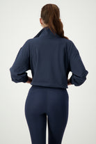 A person with long brown hair tied in a ponytail is standing with their back facing the camera. They are wearing a DRIVEN Gym Apparel Lara Half Zip Jacket (navy) with the collar turned up and form-fitting navy leggings, embodying athleisure style. The background is plain and light-colored.