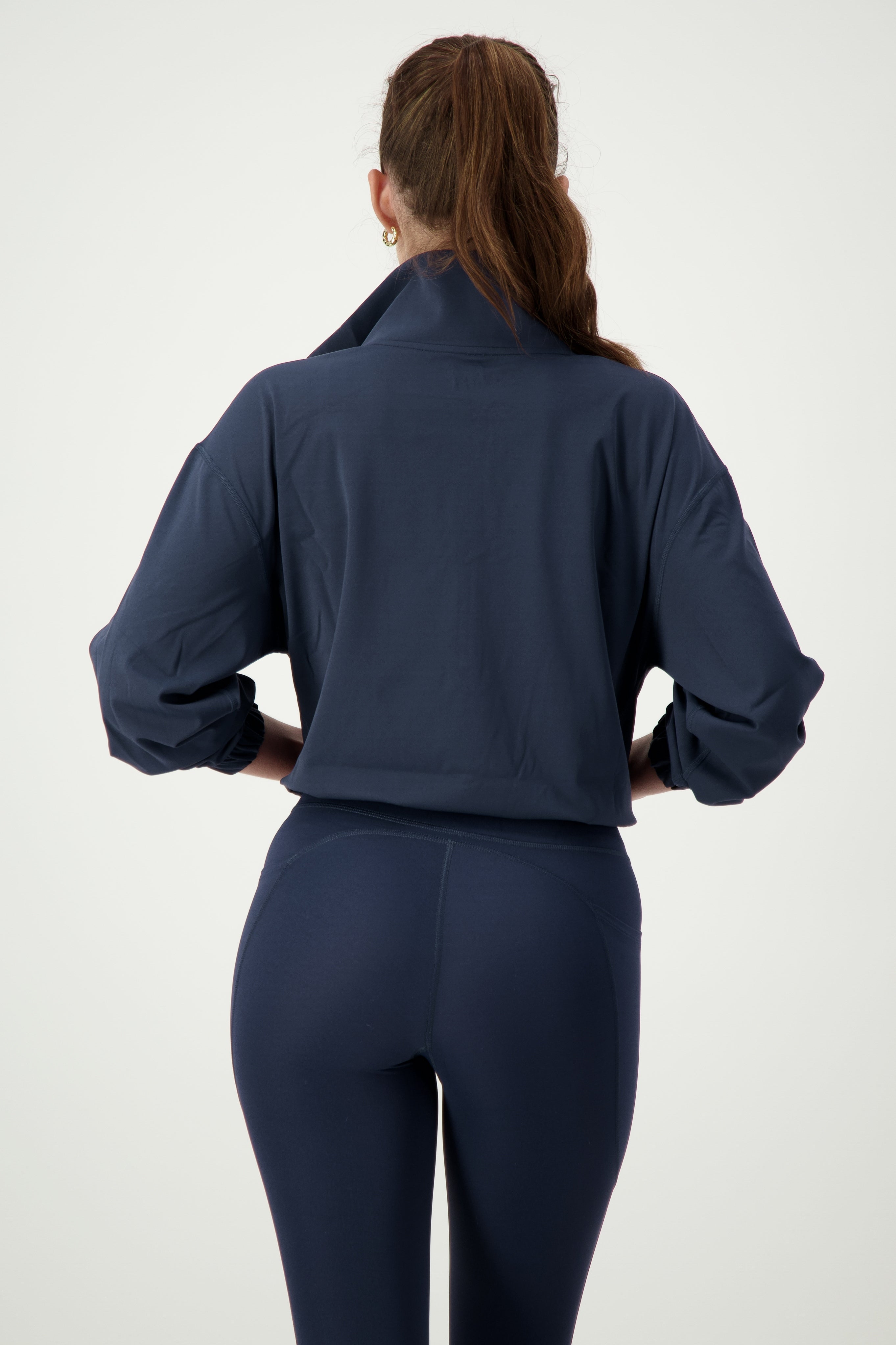 A person with long brown hair tied in a ponytail is standing with their back facing the camera. They are wearing a DRIVEN Gym Apparel Lara Half Zip Jacket (navy) with the collar turned up and form-fitting navy leggings, embodying athleisure style. The background is plain and light-colored.
