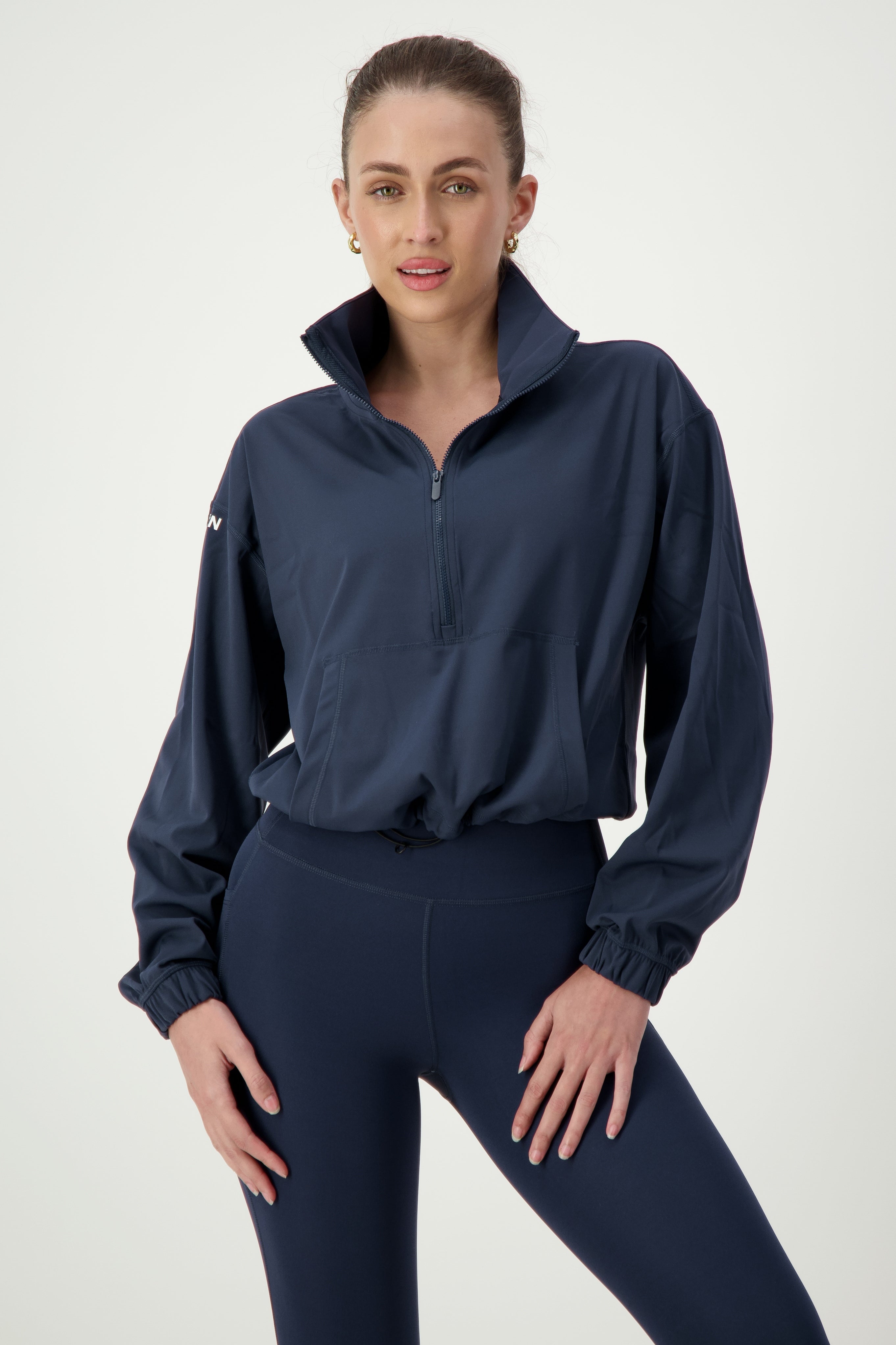 A woman wearing a navy blue Lara Half Zip Jacket (navy) from DRIVEN Gym Apparel and matching high-waisted leggings, perfect examples of stylish activewear, stands with one hand on her hip. She has her hair tied back and is smiling slightly. The jacket has a half-zip front and elasticized cuffs. The background is plain and light.