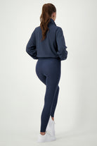 A woman with a ponytail is facing away and wearing a dark blue Lara Half Zip Jacket (navy) from DRIVEN Gym Apparel and matching leggings, showcasing her stylish fitness apparel. She stands against a plain white background, with white sneakers completing her gym wear ensemble.