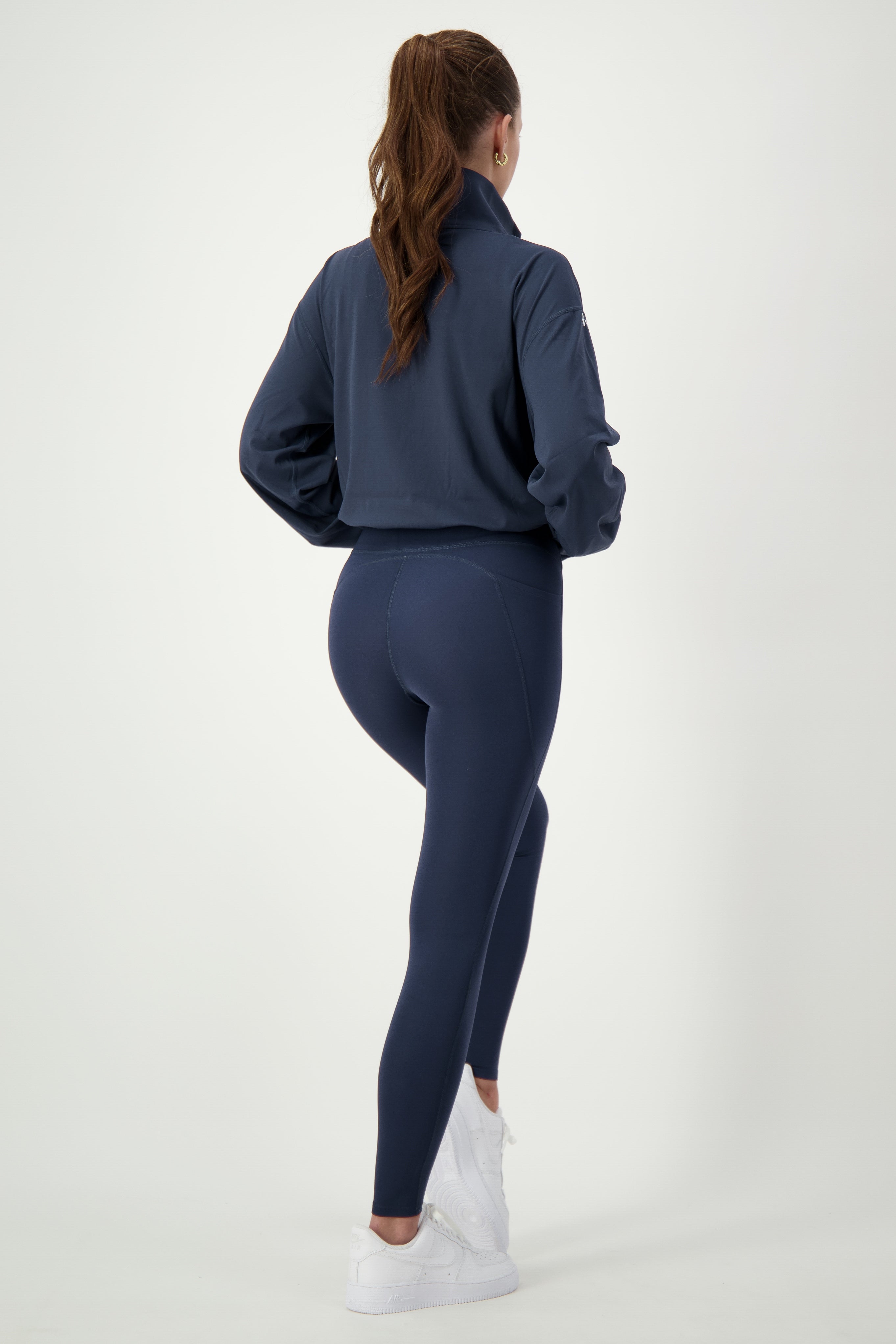 A woman with a ponytail is facing away and wearing a dark blue Lara Half Zip Jacket (navy) from DRIVEN Gym Apparel and matching leggings, showcasing her stylish fitness apparel. She stands against a plain white background, with white sneakers completing her gym wear ensemble.