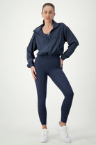 A woman stands against a plain white background, modeling gym wear. She wears a DRIVEN Gym Apparel Lara Half Zip Jacket (navy), matching high-waisted leggings, and white sneakers. Her hair is pulled back into a ponytail, and she poses with one hand on her hip and the other by her side.