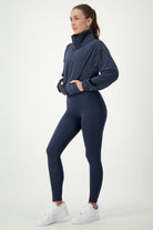 A woman stands in profile wearing a dark blue Lara Half Zip Jacket (navy) from DRIVEN Gym Apparel and matching leggings, embodying modern fitness apparel. Her hands are tucked into the pockets of the jacket, and she sports white sneakers. With long hair tied back against a plain white background, she showcases stylish activewear for every fitness enthusiast.
