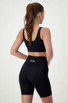 A person wearing a black Luna V-Neck Top from DRIVEN Gym Apparel and black high-waisted shorts stands with their back to the camera. Their hair is tied in a high ponytail, and they place their hands on their waist. The fitness apparel shorts have the word "DRIVEN" written on the waistband.