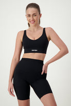 A woman with long hair is smiling and posing in black fitness apparel, including a Luna V-Neck Top (black) from DRIVEN Gym Apparel and high-waisted shorts. She has one hand on her hip against a plain light background.
