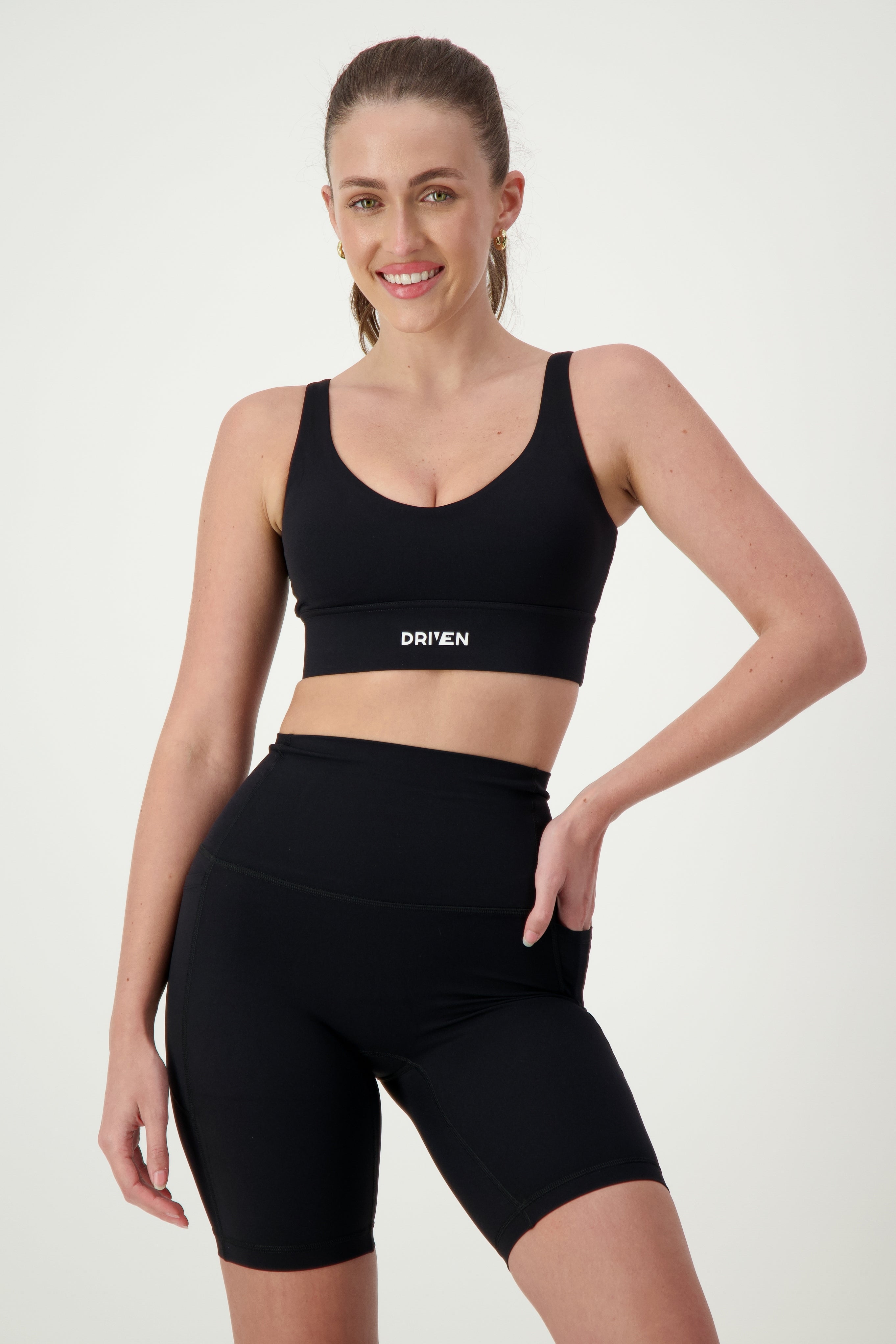 A woman with long hair is smiling and posing in black fitness apparel, including a Luna V-Neck Top (black) from DRIVEN Gym Apparel and high-waisted shorts. She has one hand on her hip against a plain light background.