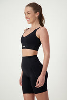 A woman with long hair tied back in a ponytail is wearing sleek fitness apparel: a black sports bra and high-waisted black bike shorts. She is standing sideways, smiling, and looking forward. The background is plain white.