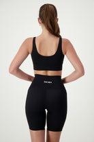 A woman stands facing away, wearing a Luna V-Neck Top (black) and high-waisted black athletic shorts with the word "DRIVEN" on the waistband. Embracing athleisure style, she has long hair pulled into a ponytail and her arms relaxed by her sides. The light-colored background emphasizes her chic fitness apparel from DRIVEN Gym Apparel.