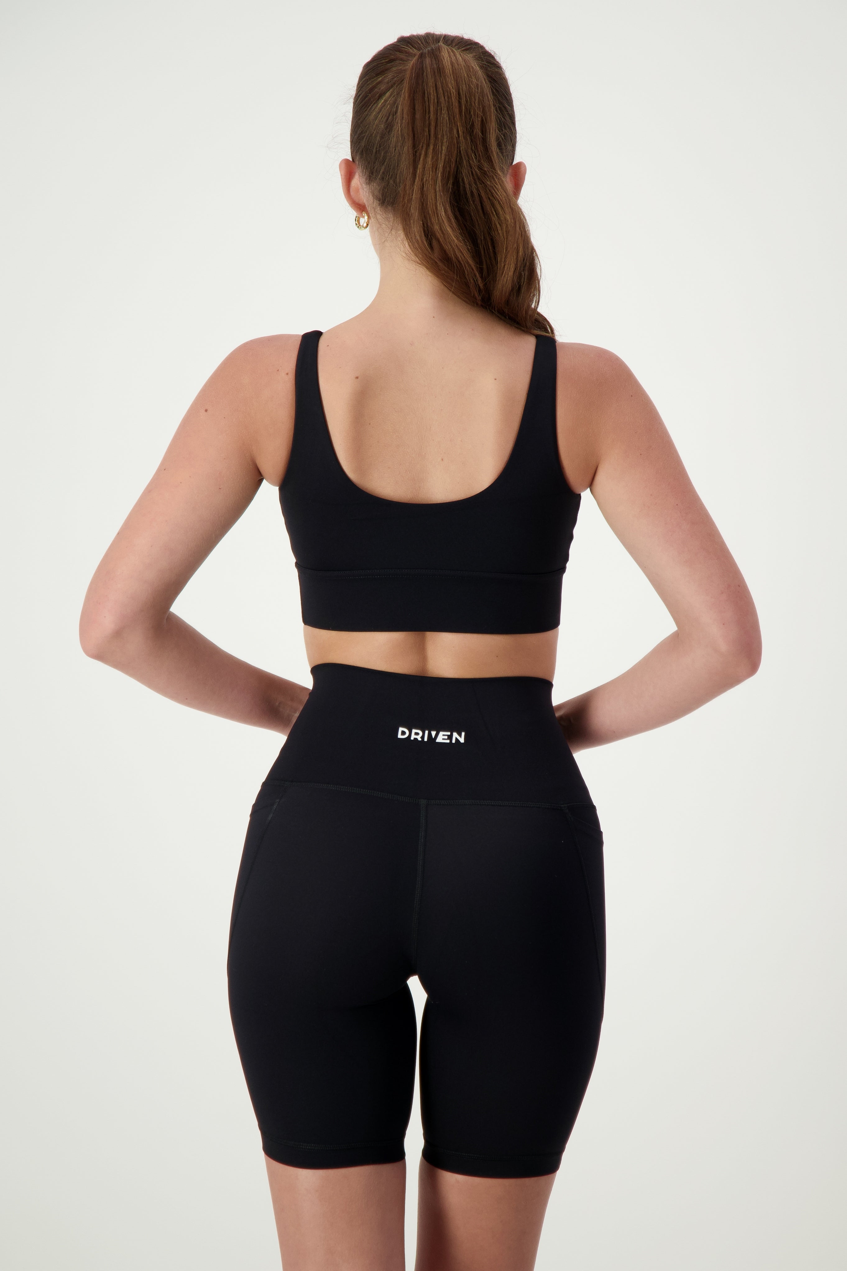 A woman stands facing away, wearing a Luna V-Neck Top (black) and high-waisted black athletic shorts with the word "DRIVEN" on the waistband. Embracing athleisure style, she has long hair pulled into a ponytail and her arms relaxed by her sides. The light-colored background emphasizes her chic fitness apparel from DRIVEN Gym Apparel.