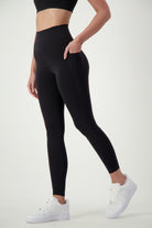 A person stands against a plain white background wearing black fitness apparel—specifically, a sports bra and DRIVEN Gym Apparel Phone Pocket Tights (black)—paired with white sneakers. The person’s left hand is placed in the pocket of the leggings, and only their upper legs and part of their torso are visible.