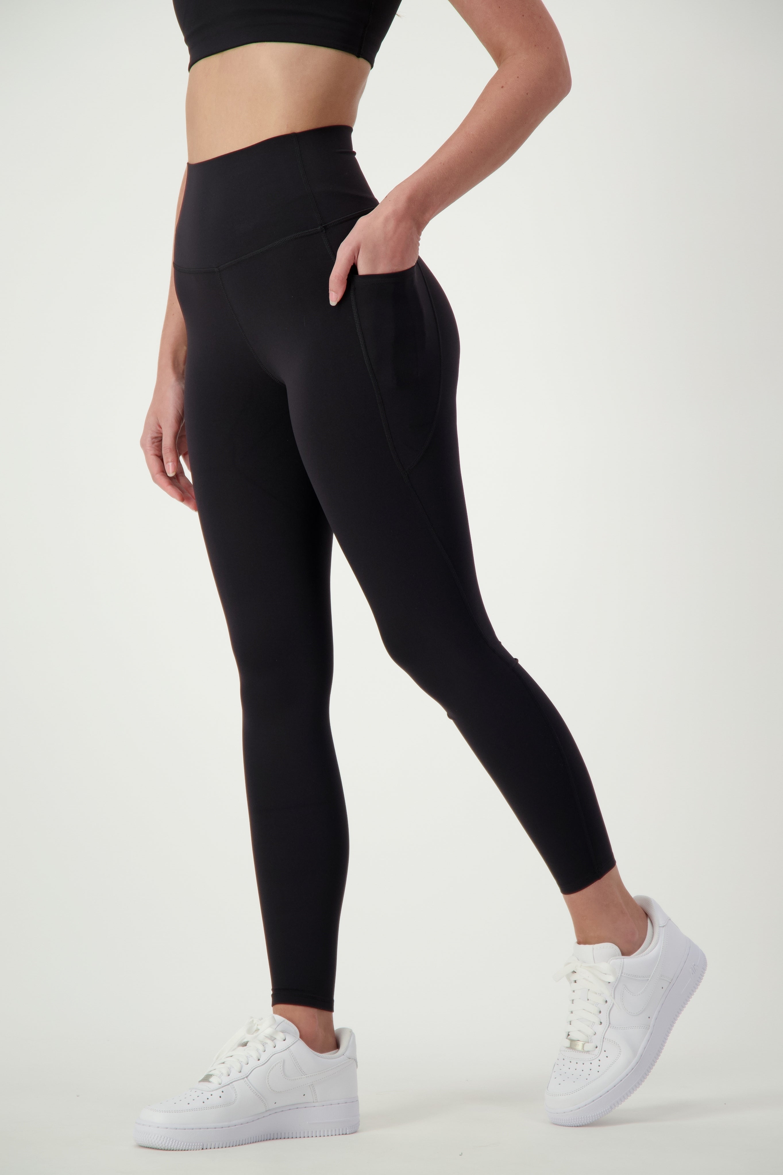 A person stands against a plain white background wearing black fitness apparel—specifically, a sports bra and DRIVEN Gym Apparel Phone Pocket Tights (black)—paired with white sneakers. The person’s left hand is placed in the pocket of the leggings, and only their upper legs and part of their torso are visible.