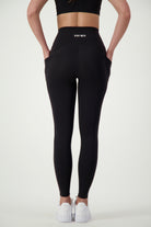 A person in black Phone Pocket Tights (black) from DRIVEN Gym Apparel with "DRIVEN" printed on the back, complemented by a black crop top and white sneakers, stands with their back facing the camera. Their chic athleisure look is set against a plain white background, showcasing stylish and functional gym wear.