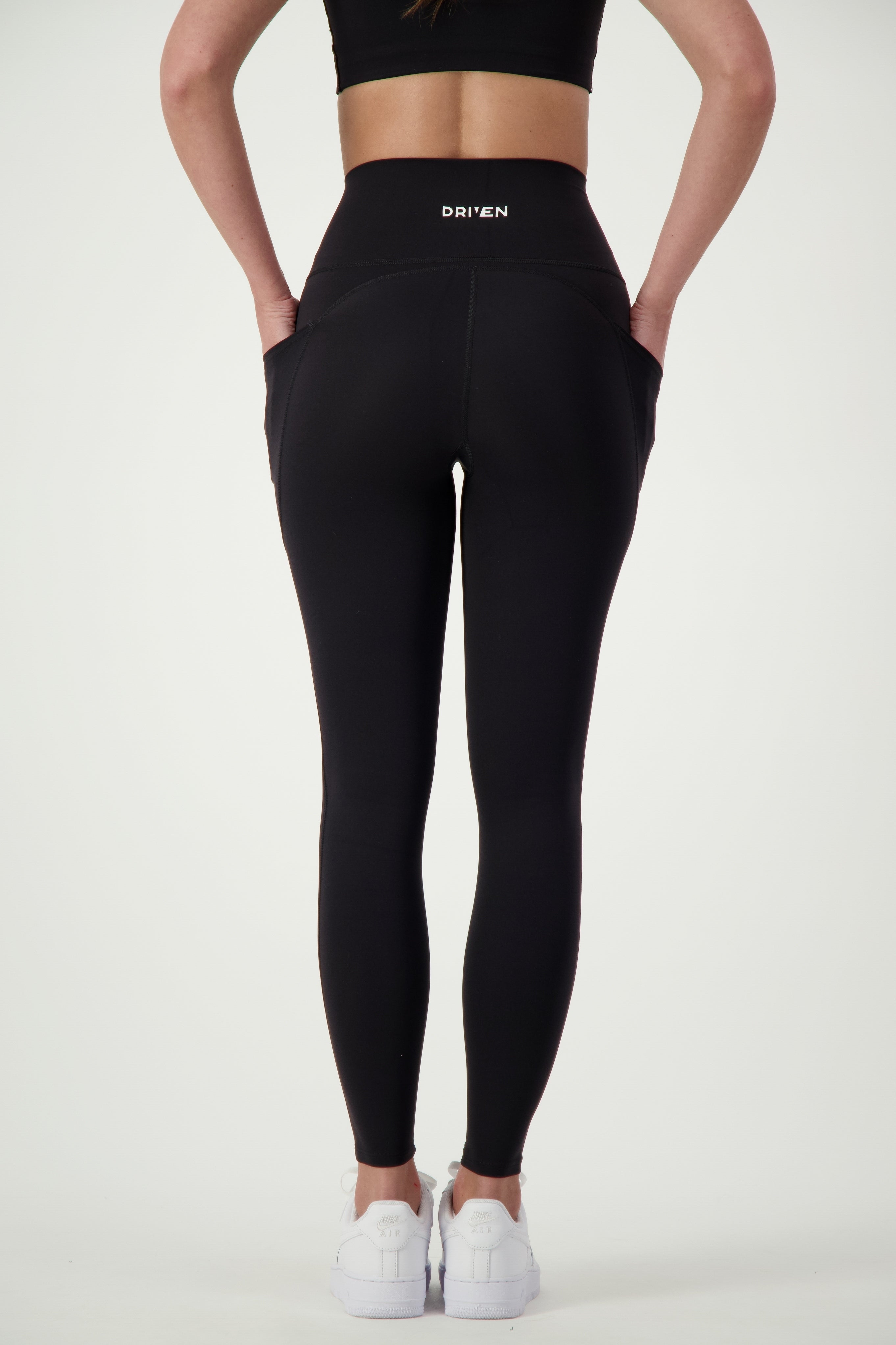 A person in black Phone Pocket Tights (black) from DRIVEN Gym Apparel with "DRIVEN" printed on the back, complemented by a black crop top and white sneakers, stands with their back facing the camera. Their chic athleisure look is set against a plain white background, showcasing stylish and functional gym wear.