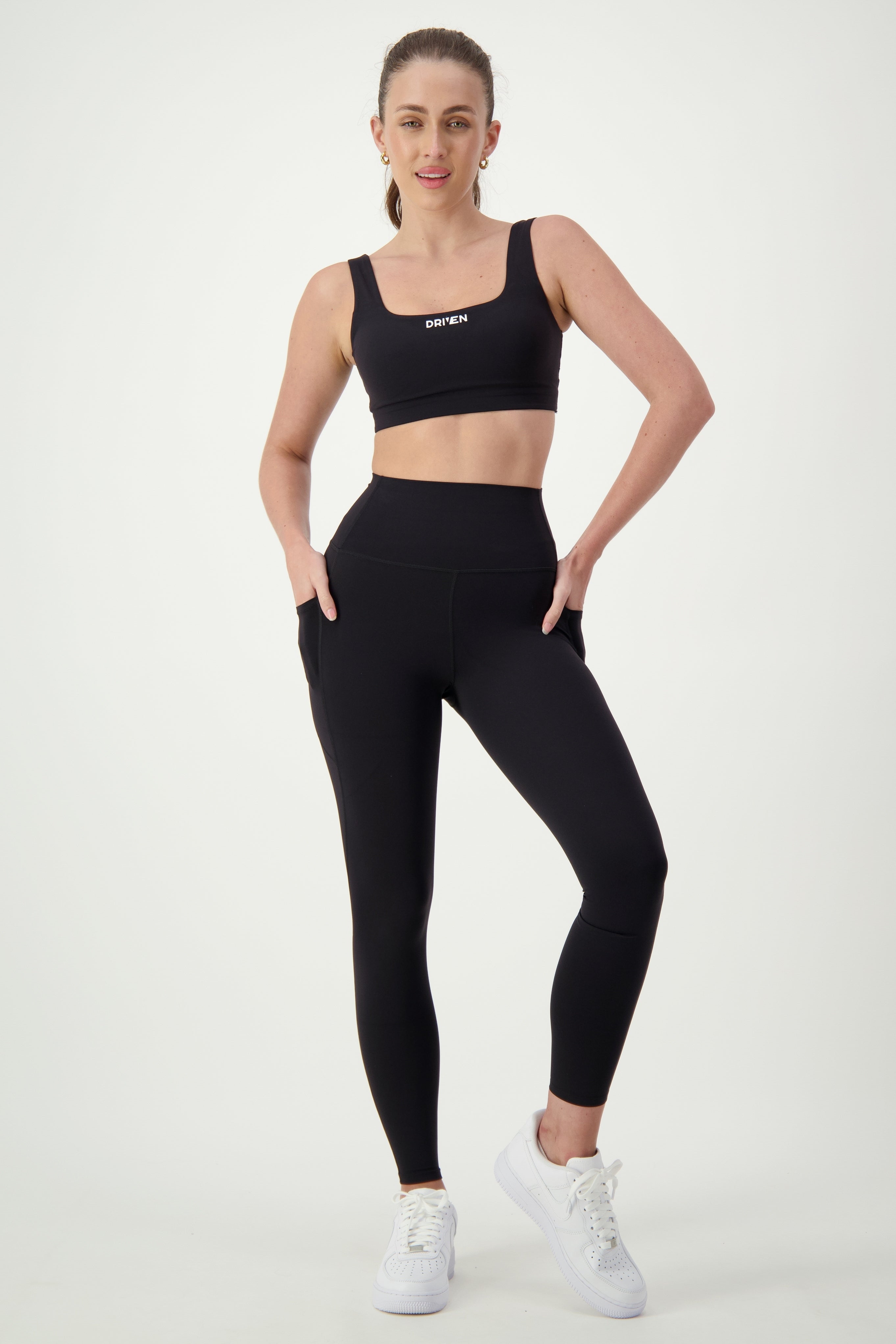 A woman is posing against a white background, wearing sleek athleisure: a black sports bra and high-waisted black DRIVEN Gym Apparel Phone Pocket Tights. She has one hand in her pocket and is wearing white sneakers. Her hair is tied back in a ponytail, and she has a confident expression.