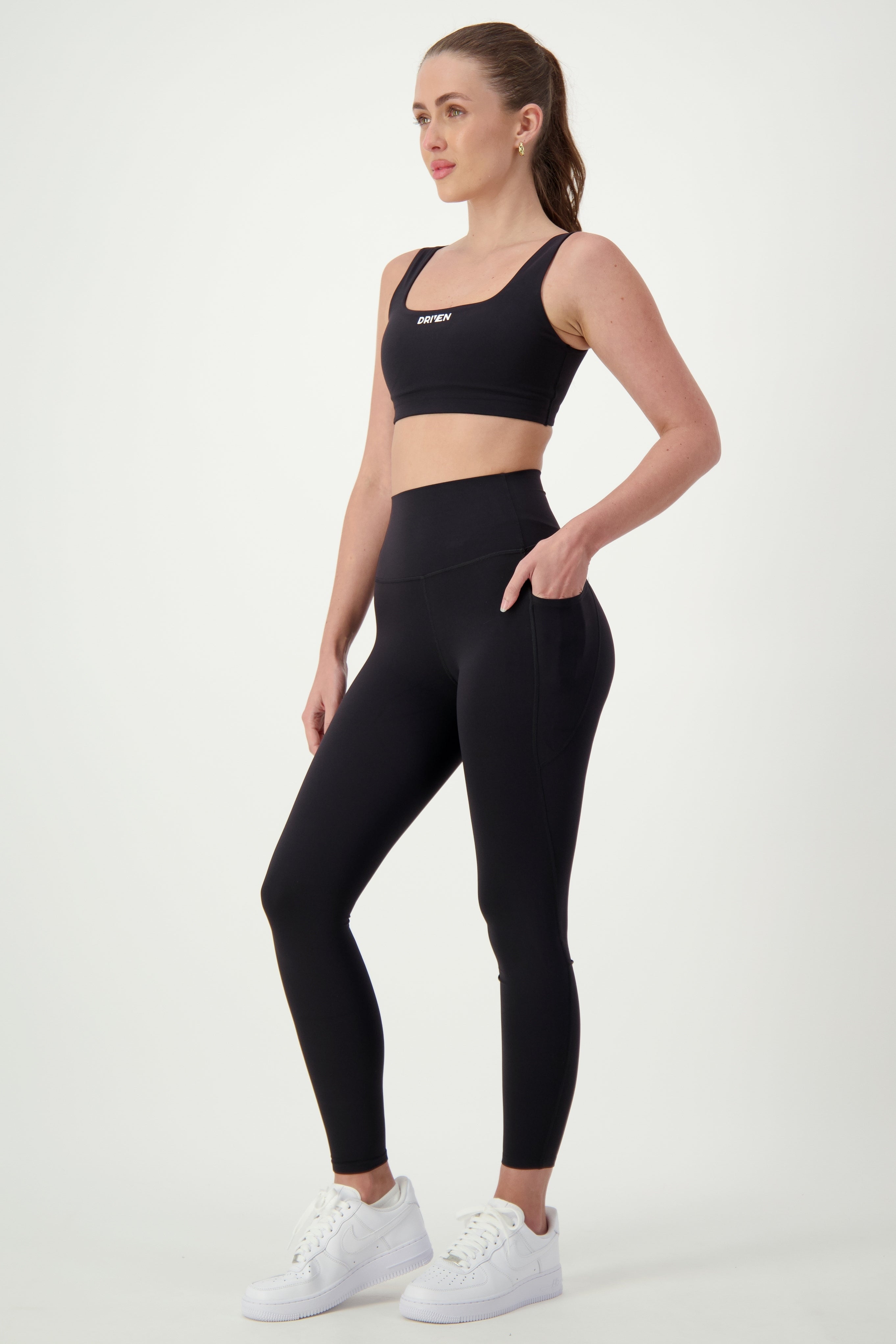 A woman stands against a plain white background, showcasing her sleek activewear. She sports a matching black sports bra and high-waisted Phone Pocket Tights (black) from DRIVEN Gym Apparel, with her hair pulled back into a ponytail and white sneakers on her feet. Posing confidently with one hand resting on her thigh, she epitomizes modern gym wear.