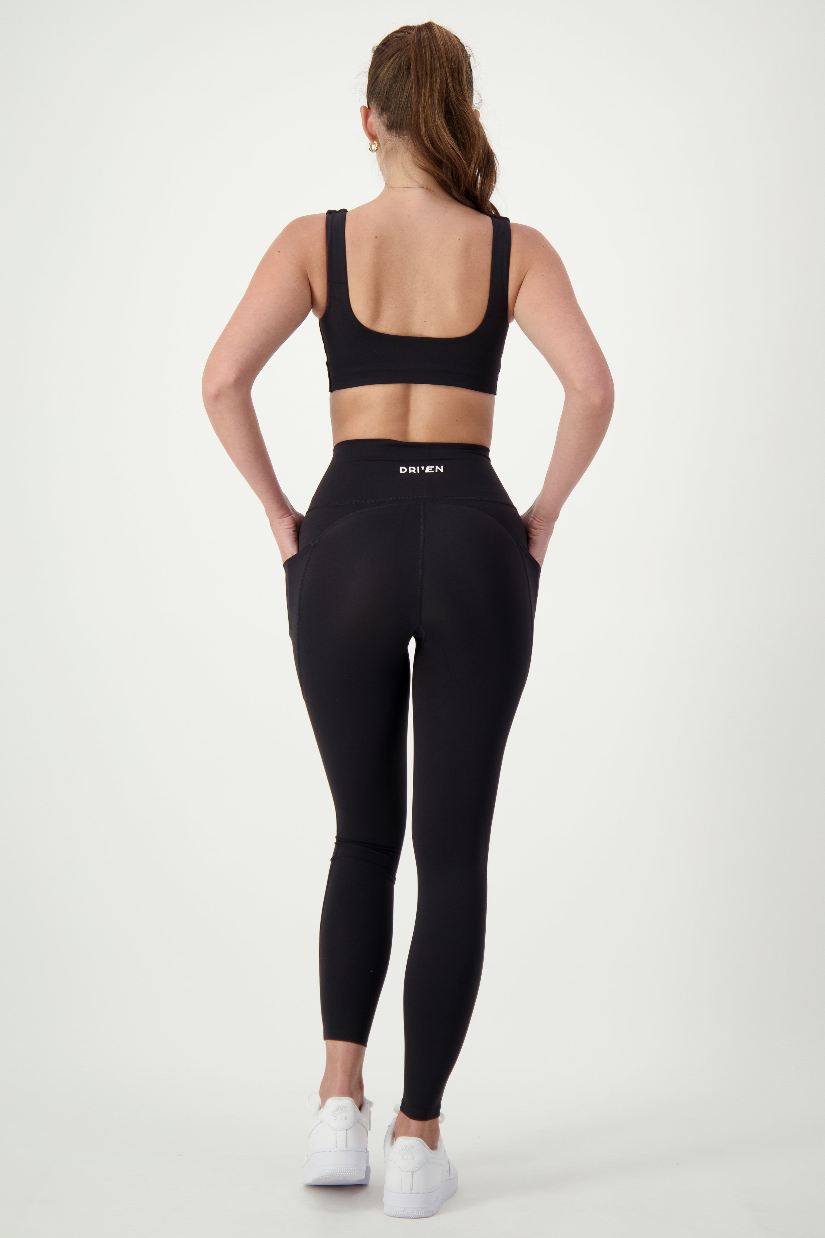A woman with long hair in a ponytail is standing with her back to the camera, showcasing chic fitness apparel. She wears a black sports bra, a pair of DRIVEN Gym Apparel Phone Pocket Tights (black), and white sneakers. Her hands rest casually in her tights' pockets against the plain white background, emphasizing modern athleisure style.