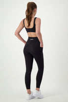 A woman with a ponytail is shown from the back, wearing black fitness apparel: a sports bra and high-waisted DRIVEN Gym Apparel Phone Pocket Tights (black). She stands against a white background, with her left hand in the pocket of her tights. She is also wearing white sneakers.