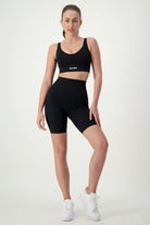 A person with long hair in a high ponytail stands against a white background, dressed in sleek black athleisure. They are wearing a sports bra with "DRIVEN" printed on the front and high-waisted Phone Pocket Bike Shorts (black) from DRIVEN Gym Apparel, paired with white sneakers.