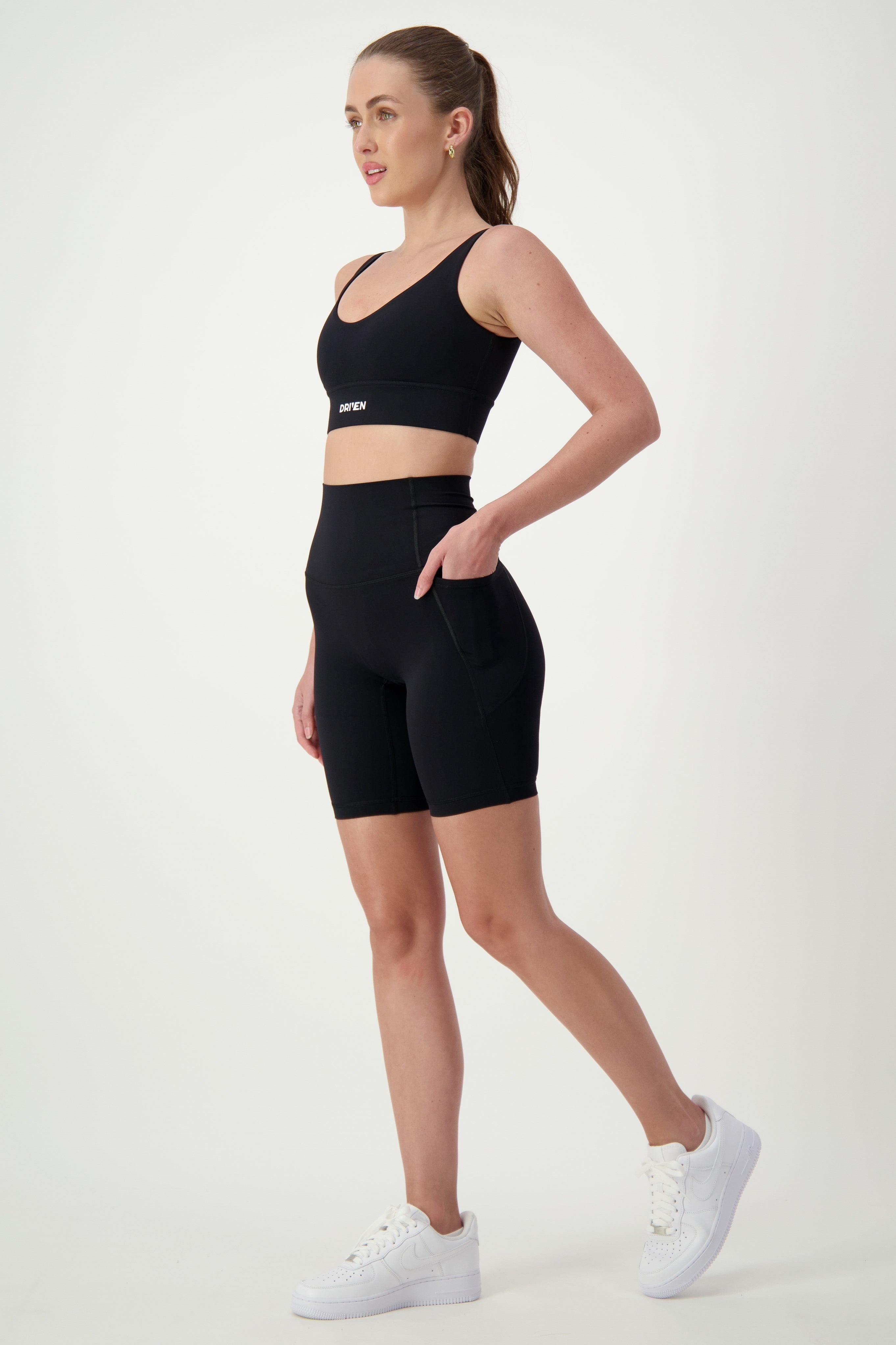 A woman dressed in black gym wear, including a sports bra and matching high-waisted Phone Pocket Bike Shorts (black) by DRIVEN Gym Apparel, stands against a plain white background. She is wearing white sneakers and has her hair tied back in a ponytail. She is gazing to her right with one hand in the pocket of her activewear shorts.