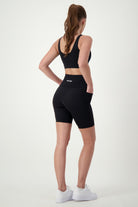 A woman with a ponytail is standing with her back to the camera, showcasing stylish activewear—a black sports bra, DRIVEN Gym Apparel's black Phone Pocket Bike Shorts, and white sneakers. Her hands are resting on her hips against a plain white background, embodying the essence of athleisure chic.