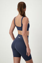 A woman with long brown hair in a ponytail is wearing matching dark blue activewear: a snug-fitting sports bra and DRIVEN Gym Apparel's Phone Pocket Bike Shorts (navy). She is standing and facing away from the camera, showcasing the back of her stylish gym wear.