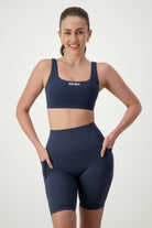 A smiling person with long hair in a ponytail is wearing matching navy blue activewear, consisting of a sports bra and high-waisted Phone Pocket Bike Shorts (navy). They stand with hands on hips against a plain background. The sports bra has the word "DRIVEN Gym Apparel" printed on the front.
