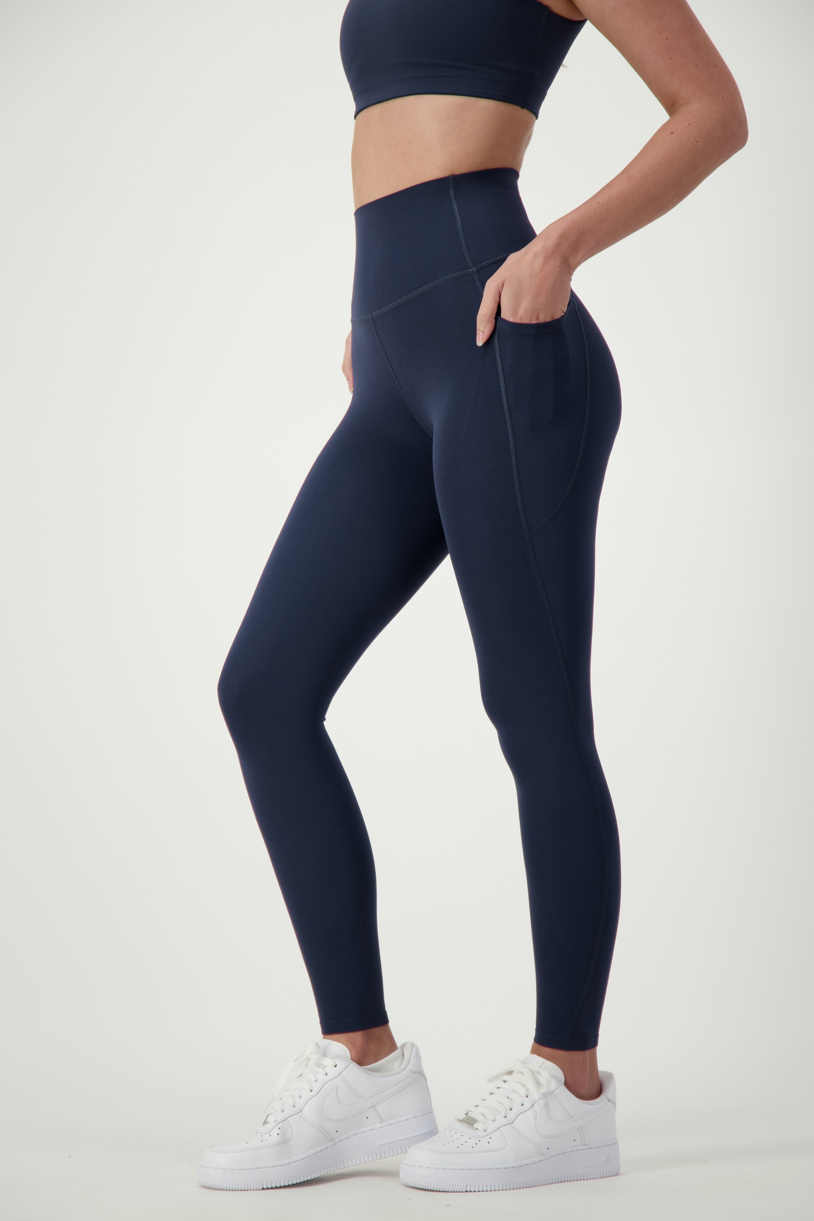 A person is wearing a blue sports bra and matching DRIVEN Gym Apparel Phone Pocket Tights (navy), epitomizing stylish activewear. They complete the look with white sneakers while standing in a neutral pose against a plain white background, one hand casually in a pocket, showcasing their midsection and legs.