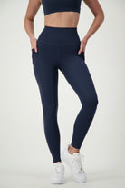 A person wearing a matching dark blue sports bra and DRIVEN Gym Apparel's Phone Pocket Tights (navy) stands against a plain white background. The individual, dressed in sleek fitness apparel, is also sporting white sneakers and posing with hands in the leggings' pockets, showcasing the workout outfit.