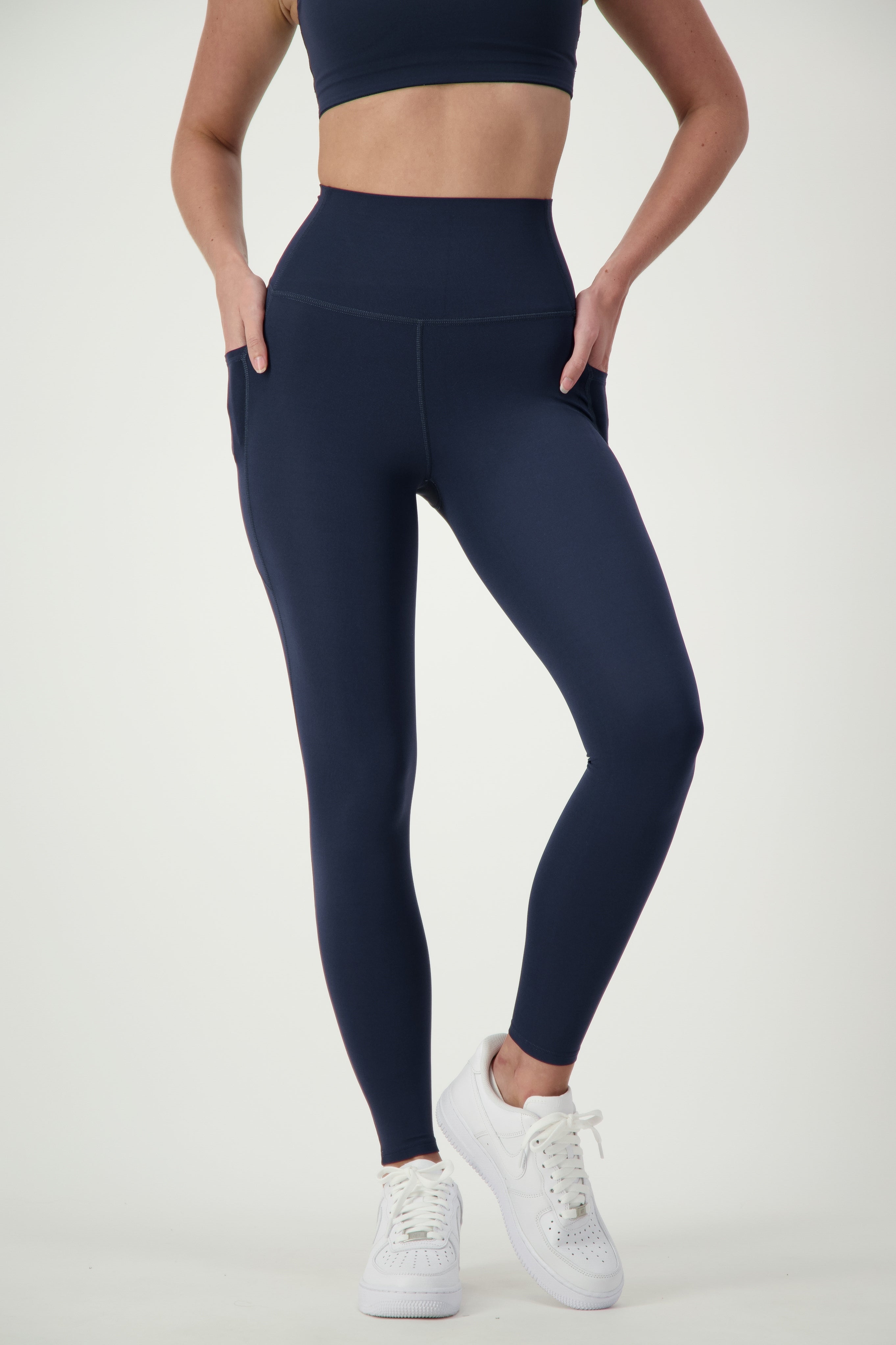 A person wearing a matching dark blue sports bra and DRIVEN Gym Apparel's Phone Pocket Tights (navy) stands against a plain white background. The individual, dressed in sleek fitness apparel, is also sporting white sneakers and posing with hands in the leggings' pockets, showcasing the workout outfit.