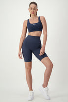 A woman poses against a plain background wearing a navy blue sports bra and matching high-waisted Phone Pocket Bike Shorts (navy), epitomizing athleisure. She has white sneakers on and a ponytail. The set features a "DRYWATE" logo on the bra from DRIVEN Gym Apparel. She stands with one leg slightly bent and her hand on her hip, sporting stylish fitness apparel.