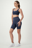 A woman stands against a plain white background wearing matching navy blue gym wear—a sports bra and DRIVEN Gym Apparel's Phone Pocket Bike Shorts (navy). She has her hair tied back in a ponytail and is wearing white sneakers. Her stance is confident, with one hand on her hip and the other resting by her side.