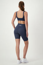A woman stands facing away, showcasing trendy gym wear. She is wearing a dark blue sports bra and high-waisted, mid-thigh length matching DRIVEN Gym Apparel Phone Pocket Bike Shorts (navy) with white sneakers. Her long hair is tied back in a ponytail. The background is plain white.