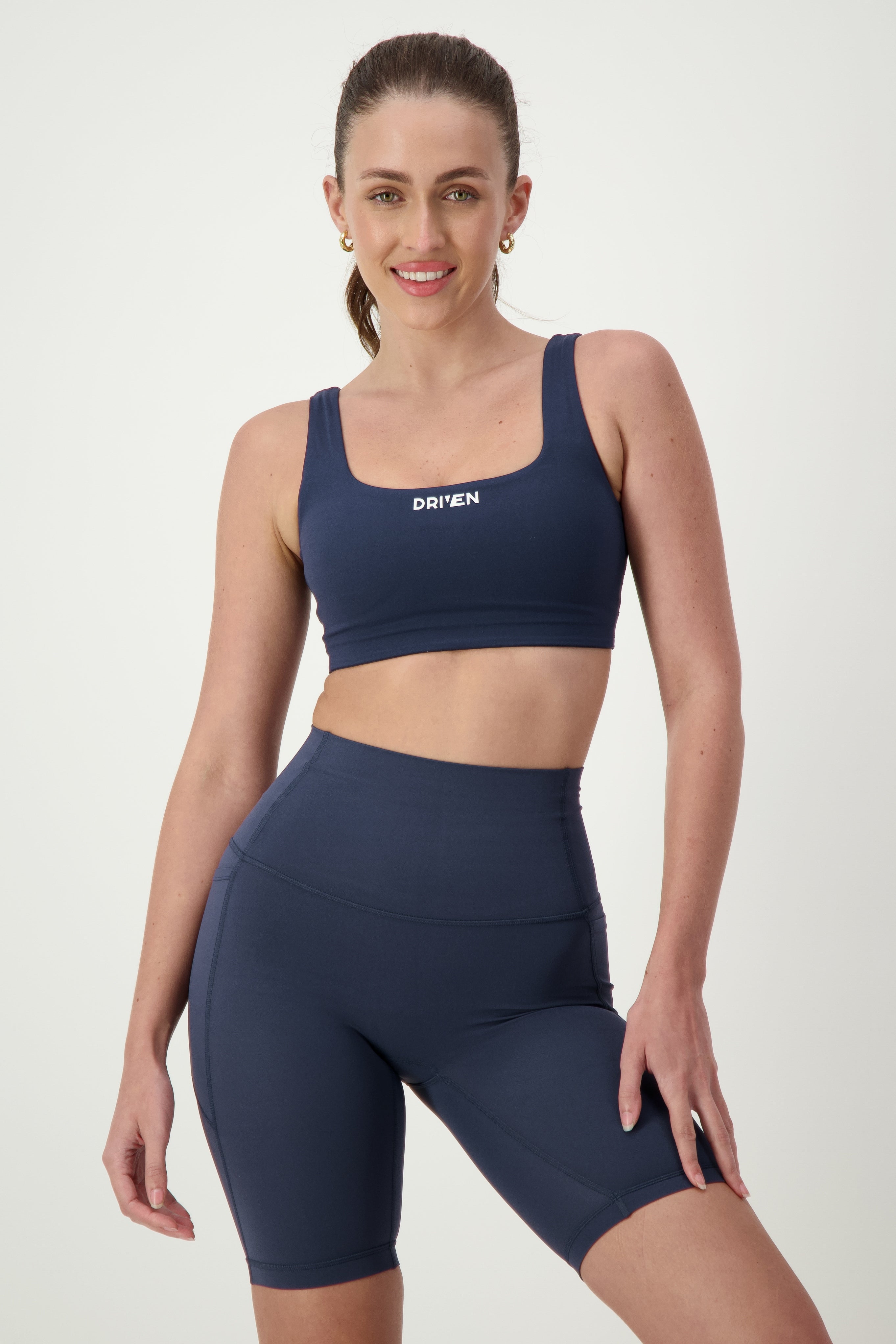 A person with long hair pulled back into a ponytail is wearing navy blue activewear: a sports bra and high-waisted matching Phone Pocket Bike Shorts (navy). The sports bra has the word "DRIVEN" written on it. Smiling, the person stands in a slight pose with one hand on their hip, showcasing their stylish gym wear from DRIVEN Gym Apparel.