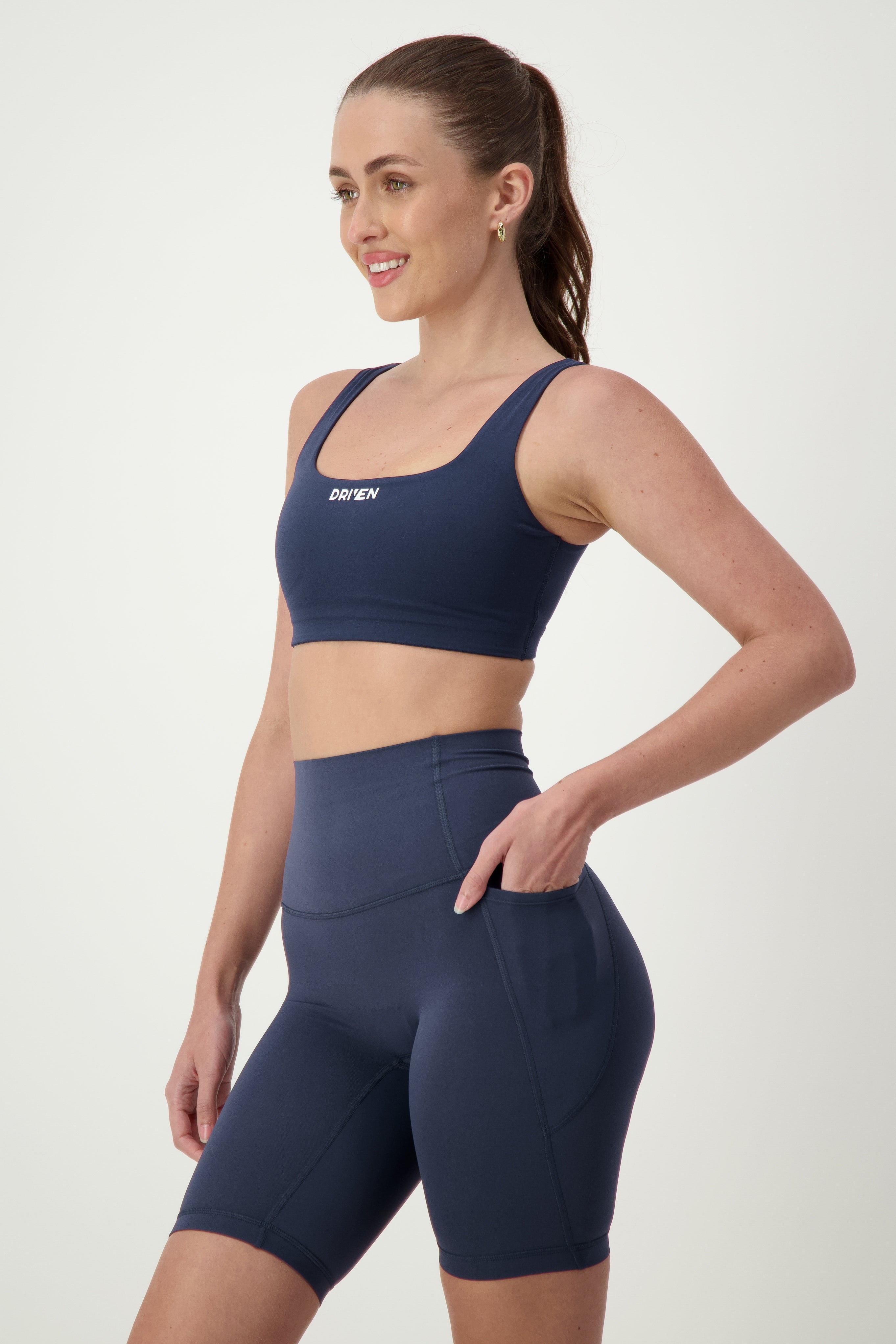 A woman is standing against a plain background, wearing a matching navy blue sports bra and high-waisted Phone Pocket Bike Shorts (navy) by DRIVEN Gym Apparel. She is smiling and looking to the side with one hand resting on her hip, and her other hand in the pocket of her athleisure wear.