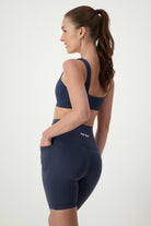 A person with long hair in a ponytail is wearing matching dark blue gym wear, consisting of a sports bra and high-waisted shorts. They are looking to the side, with one hand resting on their hip while the other is in a pocket on the side of the shorts. "DRIVEN Gym Apparel" is printed on the waistband in bold letters.