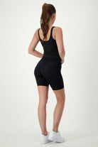 A woman with a ponytail is standing with her back to the camera, wearing black activewear: a DRIVEN Gym Apparel Ruby Staple Tank (black), cycling shorts, and white sneakers. She is posing against a plain white background.
