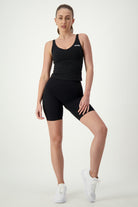 A person with long hair tied back in a ponytail stands against a plain white background wearing an all-black workout outfit. The sleek fitness apparel includes a sleeveless DRIVEN Gym Apparel Ruby Staple Tank (black) and fitted shorts. They have one leg bent slightly and are wearing white sneakers, embodying the perfect athleisure look.