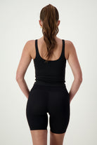 A person with long brown hair in a ponytail is standing with their back to the camera, showcasing chic athleisure. They are wearing a black Ruby Staple Tank from DRIVEN Gym Apparel and black high-waisted athletic shorts. The background is plain and light-colored, highlighting their stylish fitness apparel.