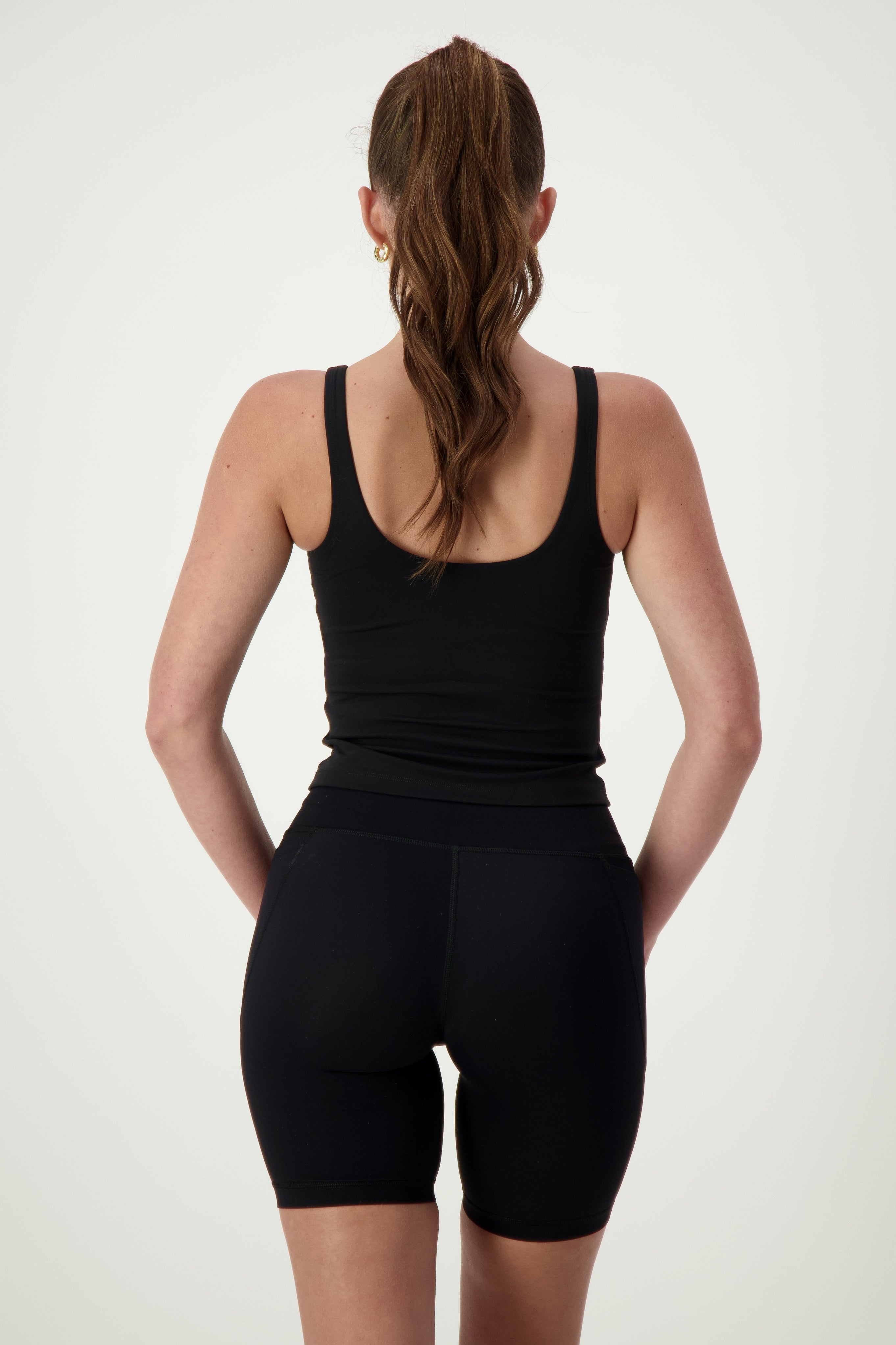 A person with long brown hair in a ponytail is standing with their back to the camera, showcasing chic athleisure. They are wearing a black Ruby Staple Tank from DRIVEN Gym Apparel and black high-waisted athletic shorts. The background is plain and light-colored, highlighting their stylish fitness apparel.
