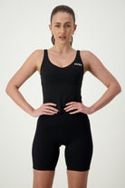 A person stands confidently with hands on hips, wearing black activewear consisting of a tank top and athletic shorts. They have a focused expression and hair pulled back in a ponytail. The Ruby Staple Tank (black) from DRIVEN Gym Apparel features the word "DRIVEN" on the chest. The background is plain white.