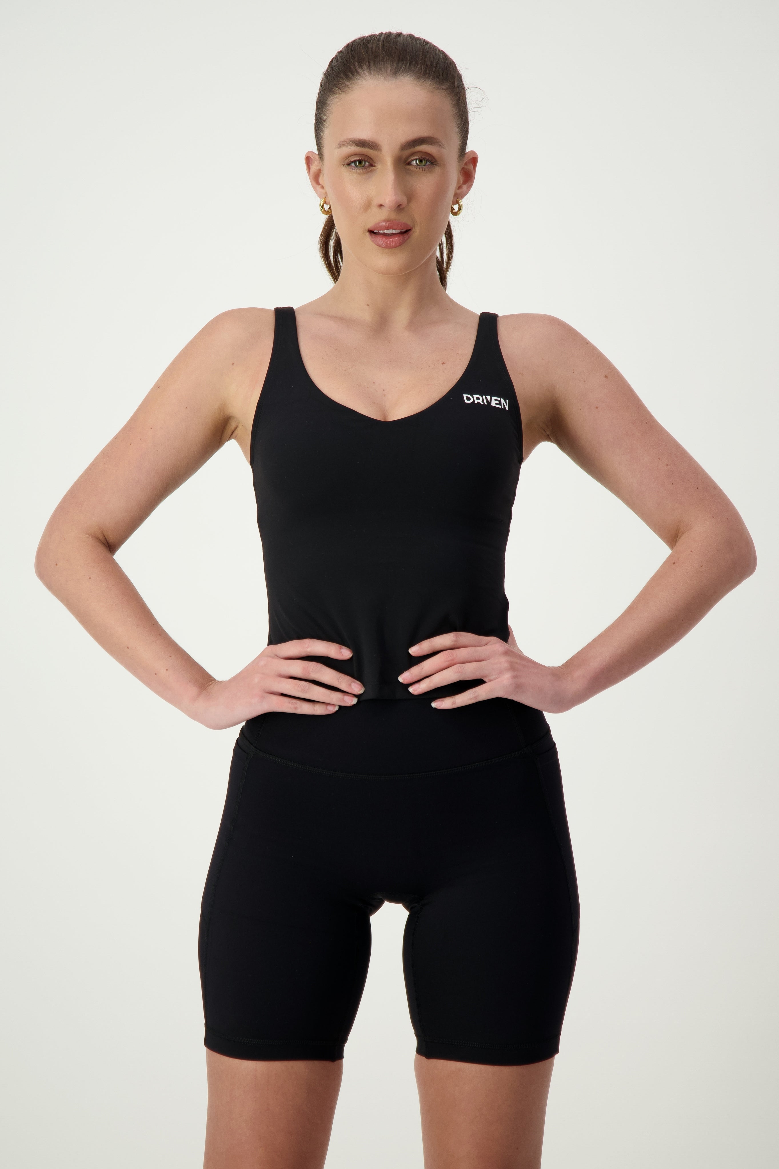 A person stands confidently with hands on hips, wearing black activewear consisting of a tank top and athletic shorts. They have a focused expression and hair pulled back in a ponytail. The Ruby Staple Tank (black) from DRIVEN Gym Apparel features the word "DRIVEN" on the chest. The background is plain white.