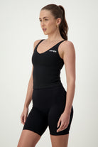 A person with long hair tied in a ponytail is wearing a black Ruby Staple Tank from DRIVEN Gym Apparel with the word "DRIVEN" on it and matching black athleisure shorts. They are standing sideways, looking into the distance. The background is plain white.